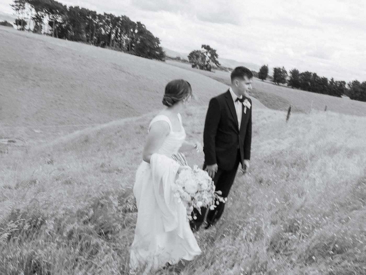 A bride and groom walking through a hillside paddock, in black and white